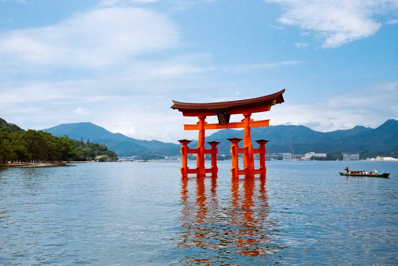 The vermilion torii gate of Itsukushima standing in the sea, reflected in the water, as a small boat of tourists rows past