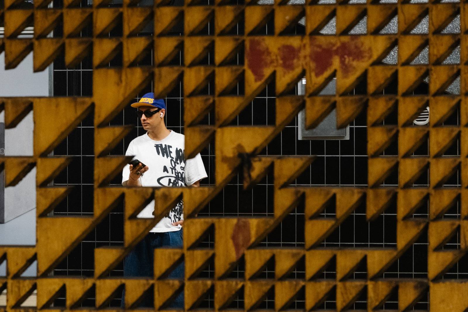 A man in a cap and sunglasses checking his phone, framed by the gaps in a rusted yellow perforated wall