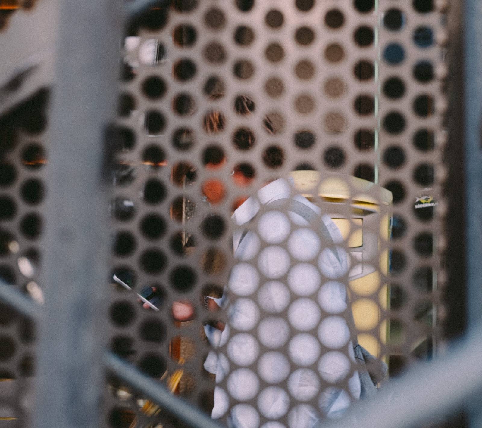 A person in a white shirt seen through the holes of a perforated metal screen, a blurred railing crossing the foreground