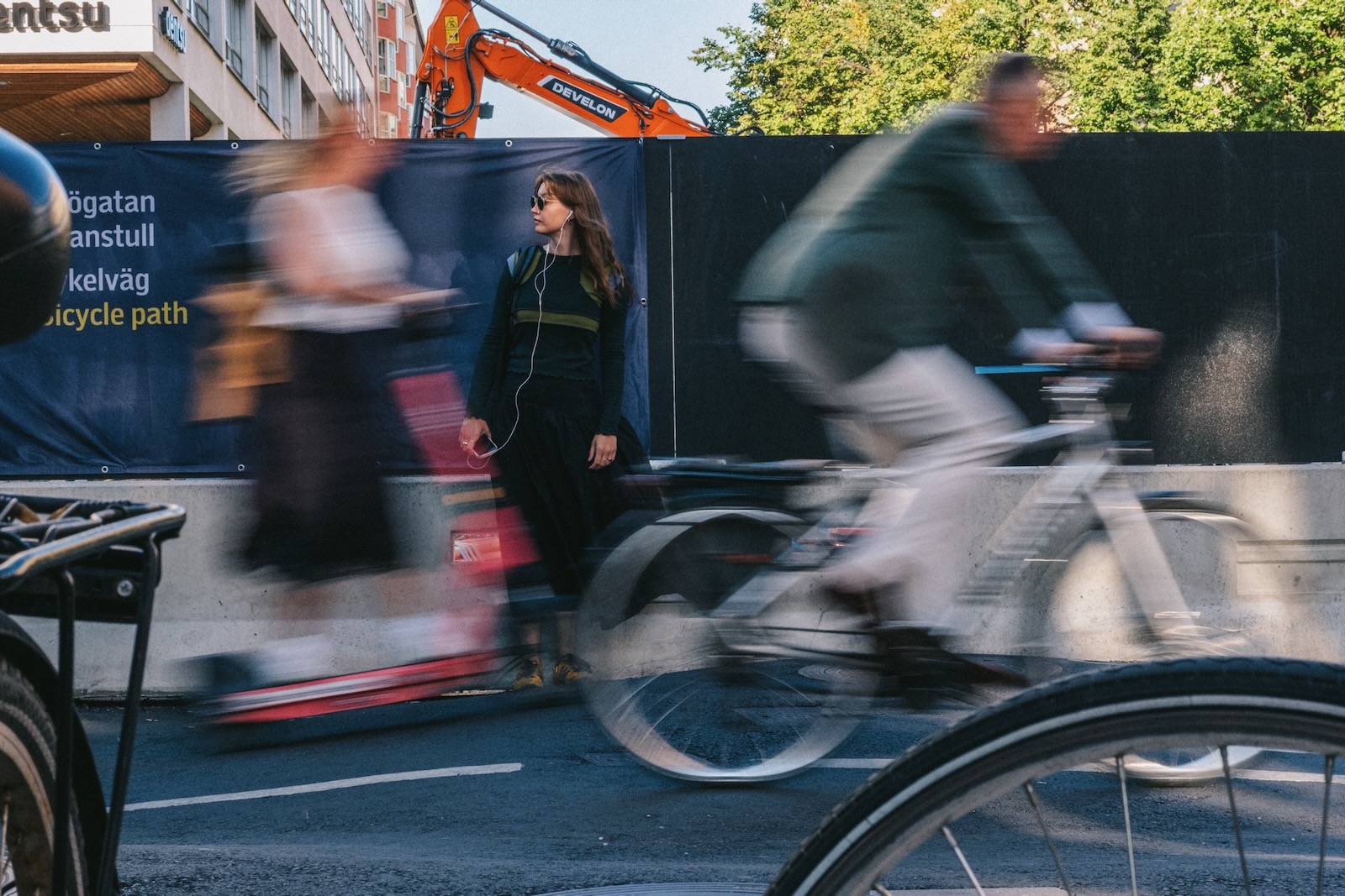 A woman stands still with earphones in while a cyclist blurs past her on a Skanstull street