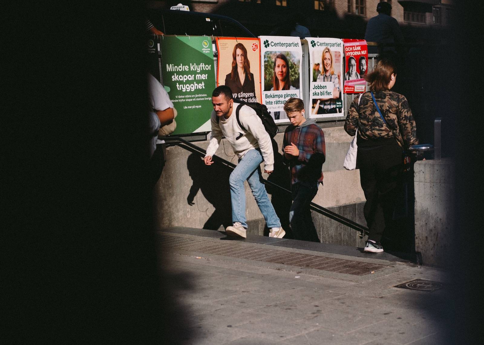 Two people climbing out of a metro entrance into low sunlight, past a wall of Swedish election posters
